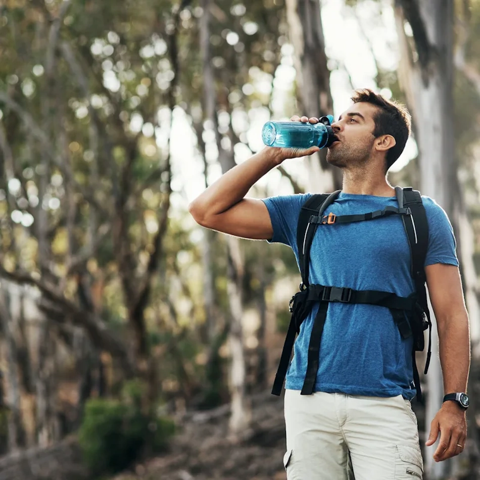 Exhausted hiker sitting in natural shade taking hydration break with water bottle during summer mountain trail hike, demonstrating heat illness prevention strategies and proper rest techniques to avoid heatstroke during outdoor adventure activities - InfoProds 2026