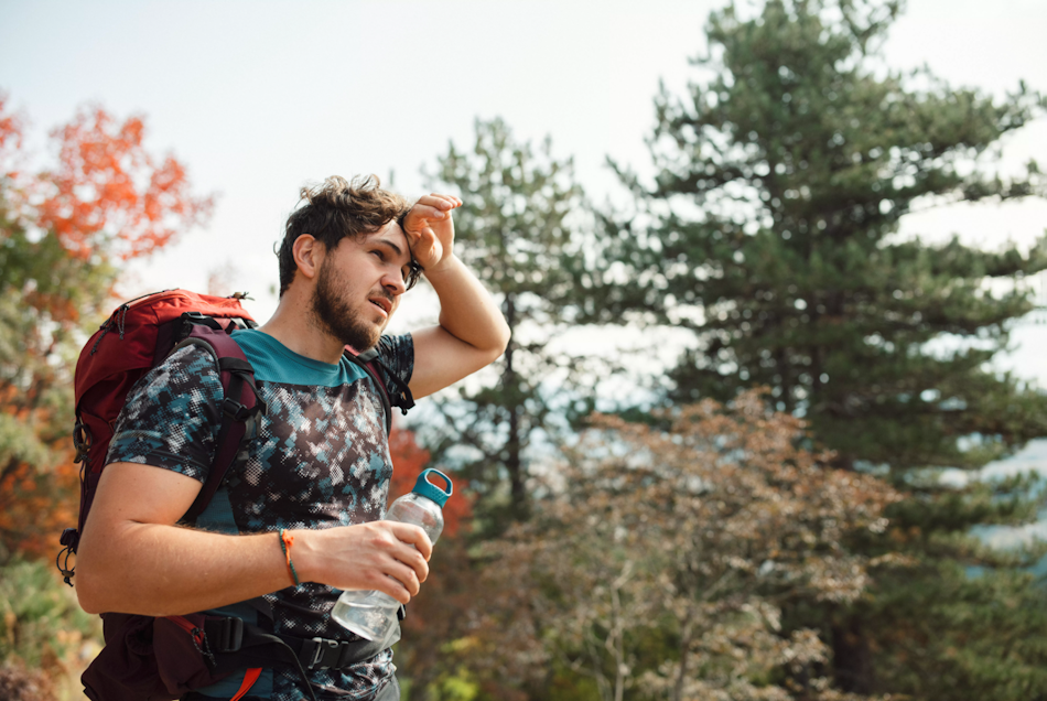 Distressed hiker showing early heat exhaustion warning signs including headache and fatigue while sitting on rocky trail during summer hike, demonstrating critical importance of recognizing heat illness symptoms before progression to dangerous heatstroke - InfoProds 2026