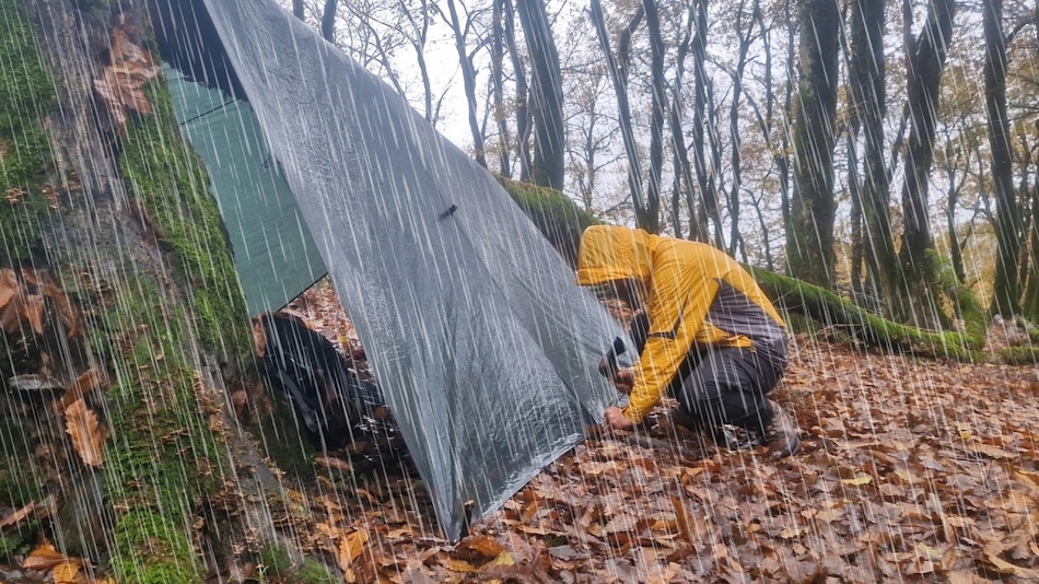 Professional rain testing facility showing three different survival shelters including tarp, tent, and bivy sack positioned under controlled irrigation system simulating heavy rainfall conditions for comparative waterproof performance analysis - InfoProds Outdoor Survival Guide 2026